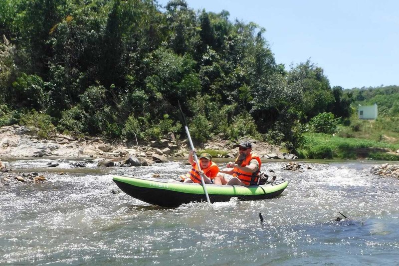 Family Kayaking Experience in Vietnam Father and son paddling together in a green inflatable kayak through shallow rocky waters.