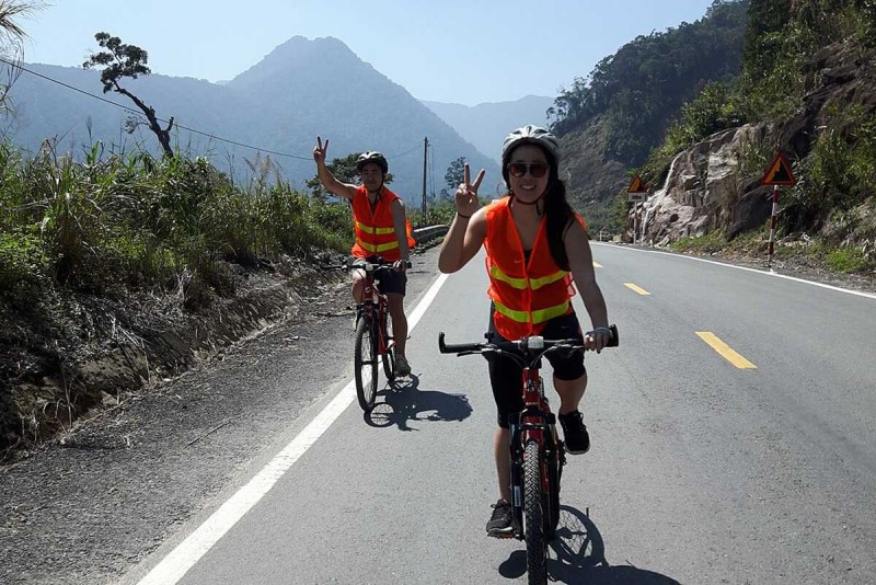 Two cyclists ride through scenic Vietnam mountains Two smiling cyclists ride uphill on a clear mountain road with high peaks in the distance