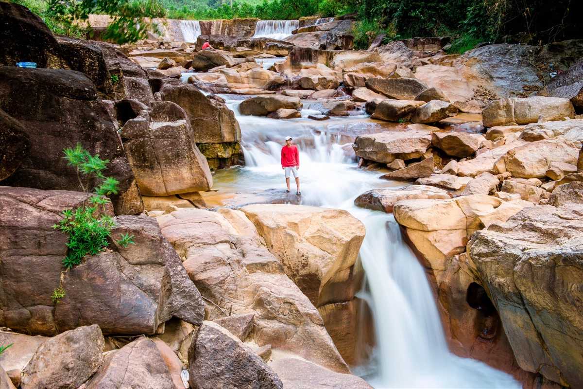 A traveler in red stands atop the rocky lower tier of Ba Ho Waterfalls with water rushing through boulders.