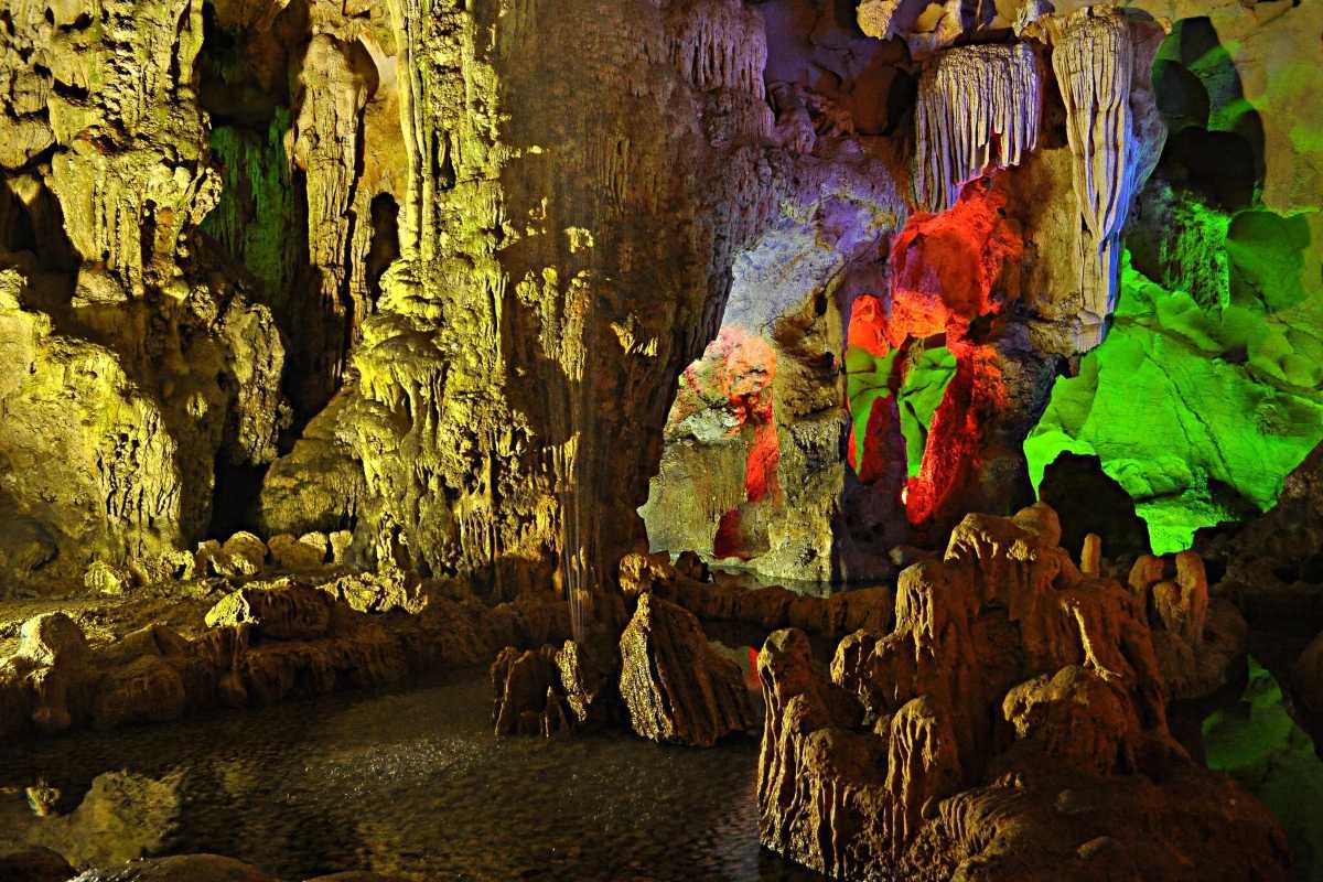 Dau Go Cave Halong Bay displays stunning stalactites illuminated by vibrant yellow, green, and red lights inside.