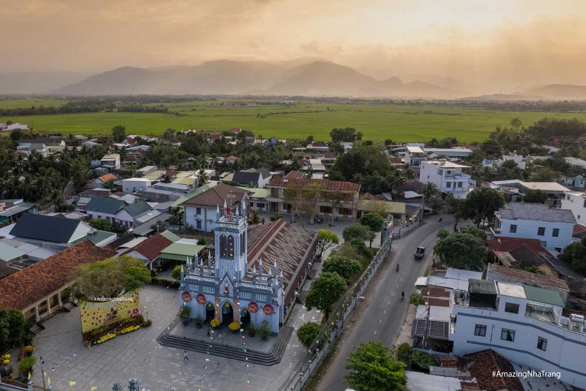 Panoramic view of Dien Khanh Town with church tower, rooftops, and green rice fields at sunset.
