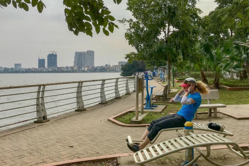 Exercise Along the Lakeside: Outdoor Gym at West Lake Hanoi A woman using an outdoor gym machine by the lakeside path at West Lake Hanoi, with city buildings in the distance.