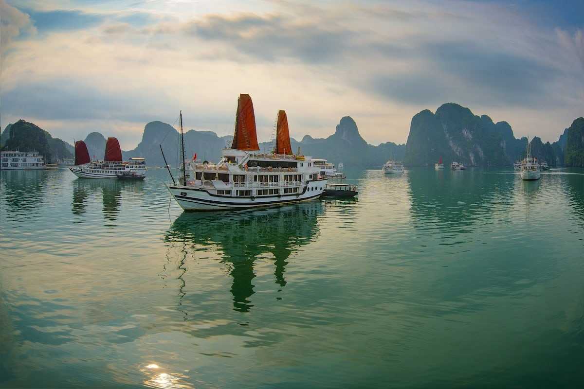 Ha Long Bay cruise boats sailing on emerald waters surrounded by towering limestone karsts under a cloudy sky.