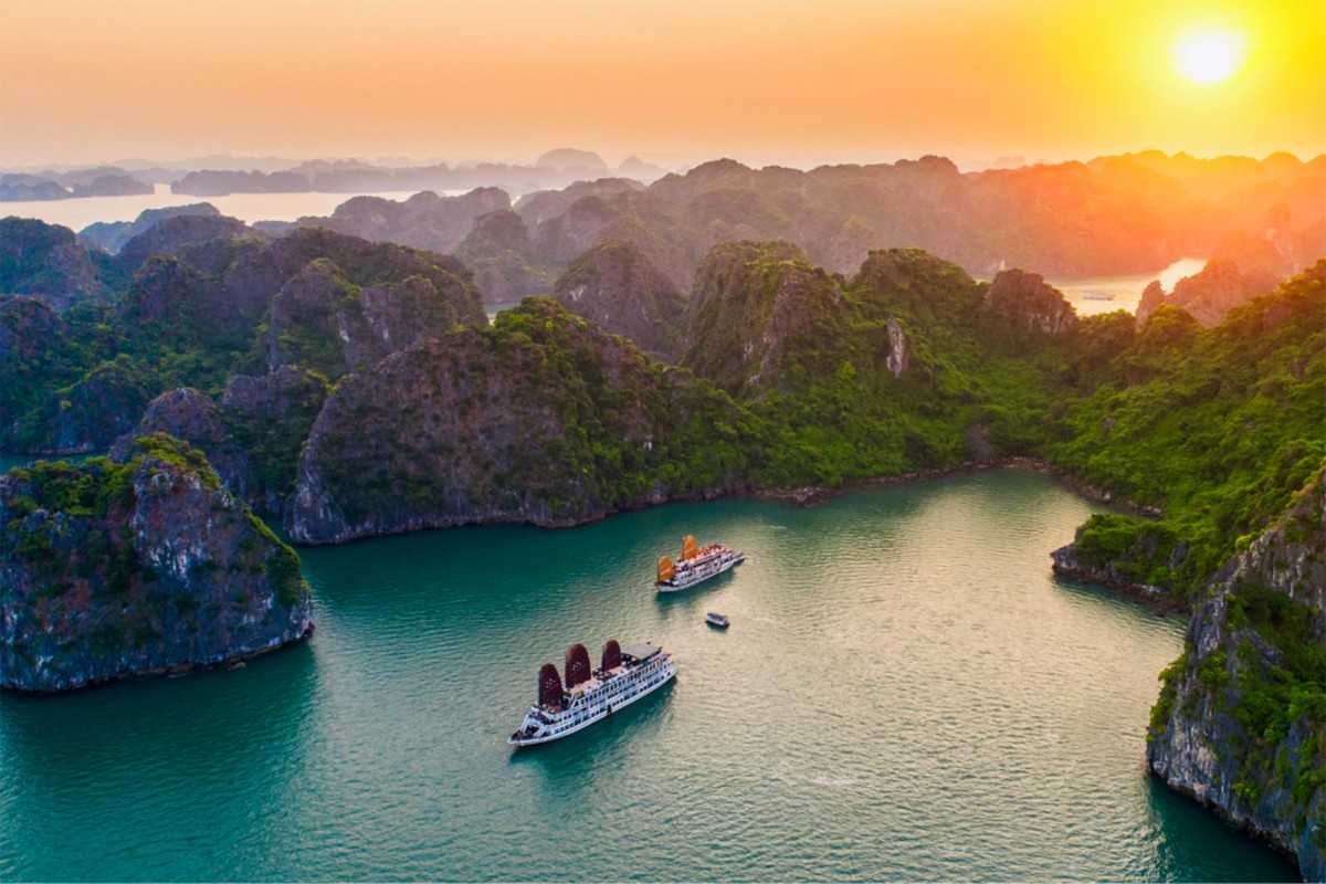 A large cruise ship with orange sails sails near limestone cliffs as the Halong Bay sunset paints the sky.
