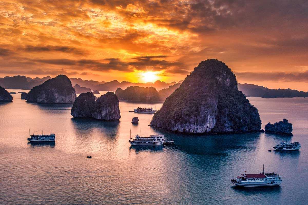 Halong Bay at sunset featuring several cruise ships surrounded by rocky islets and an orange-hued sky.