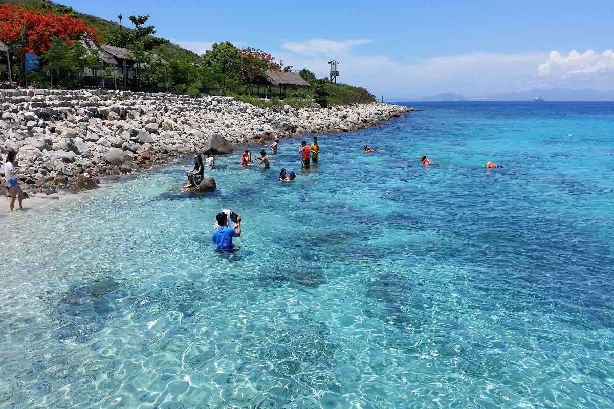 People playing and snorkeling in clear shallow waters near Hon Mun Island’s rocky shoreline.