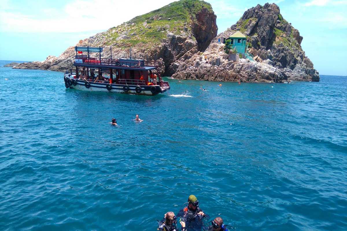 Tour boat and tourists swimming near a rocky cliff area of Hon Mun Island under bright skies.
