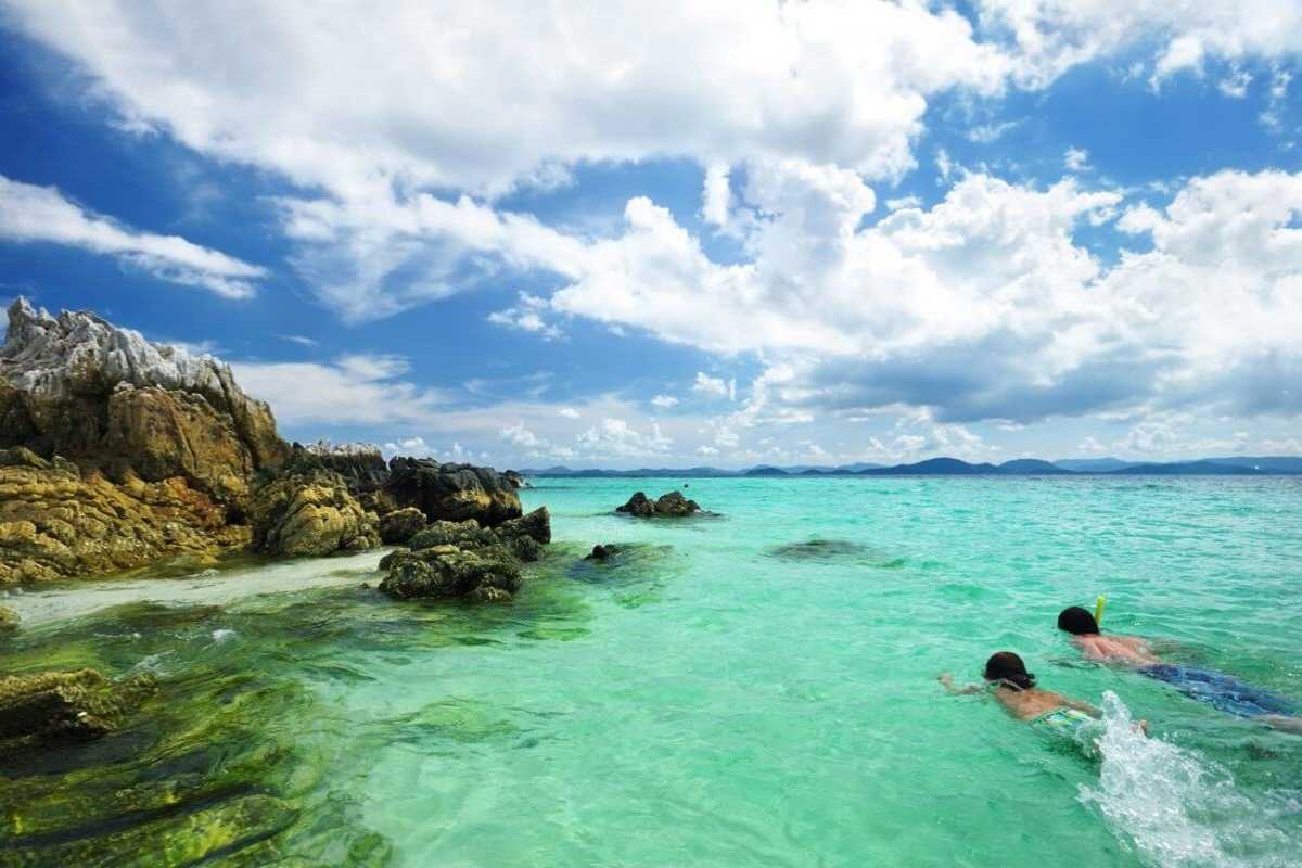 Two snorkelers in clear turquoise water near rocky outcrops of Hon Mun Island under a partly cloudy sky.