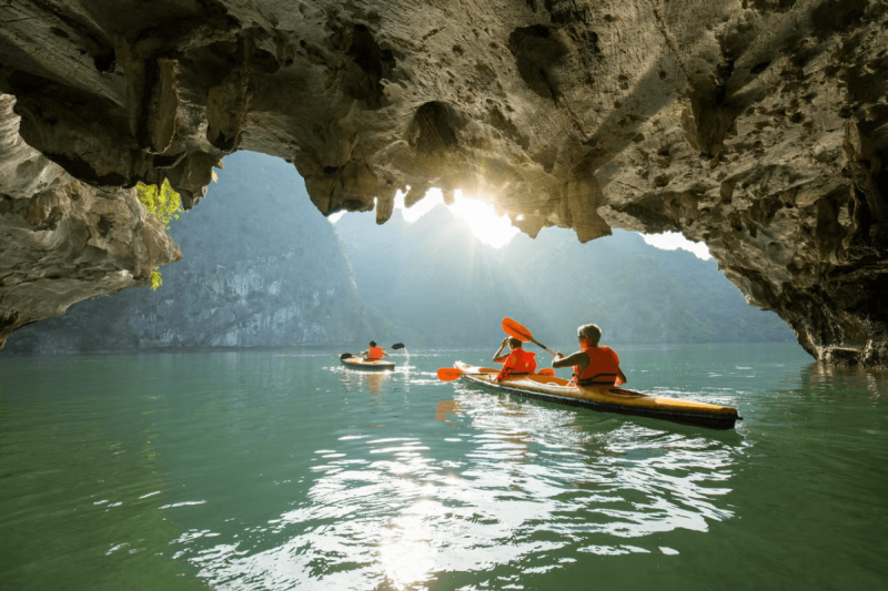 kayaking-caves-halong-bay-titov-island.png Kayakers paddling through a scenic cave in Halong Bay, with sunlight streaming through the limestone formations near Titov Island.