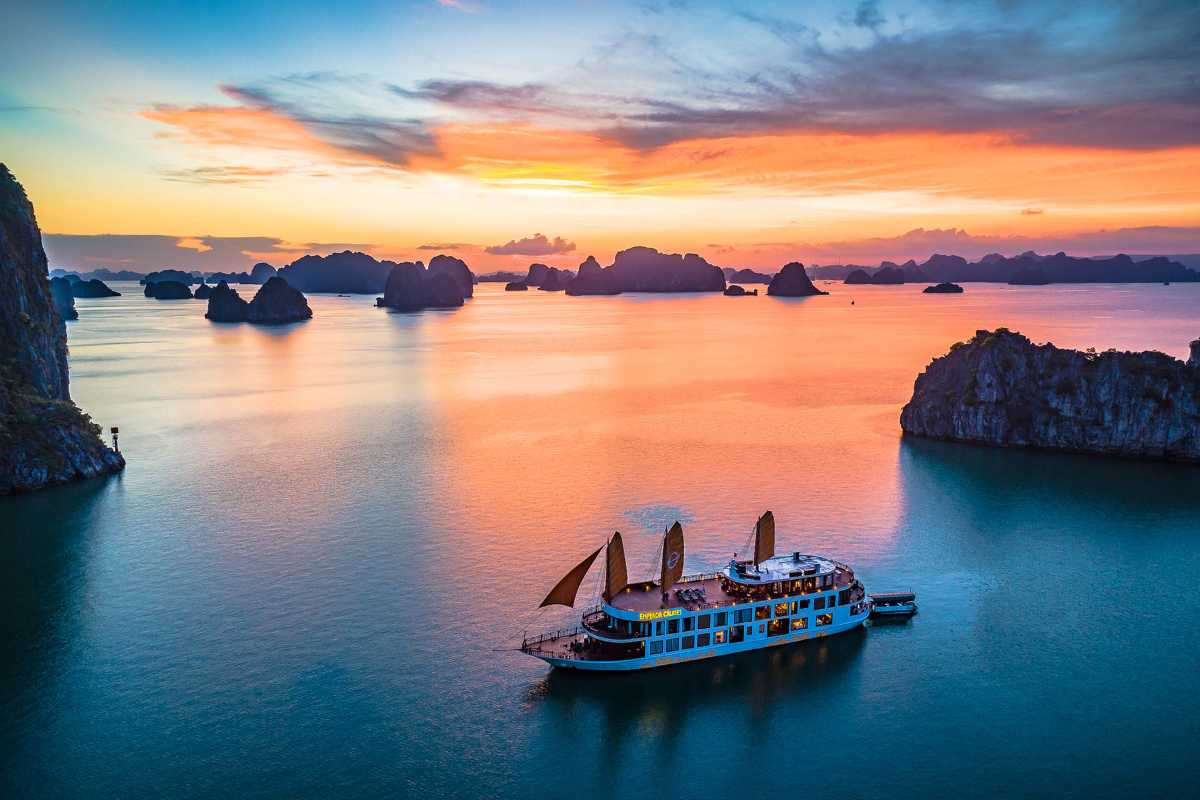 A luxury cruise with lit decks sails through Halong Bay as golden light reflects on the water and cliffs.