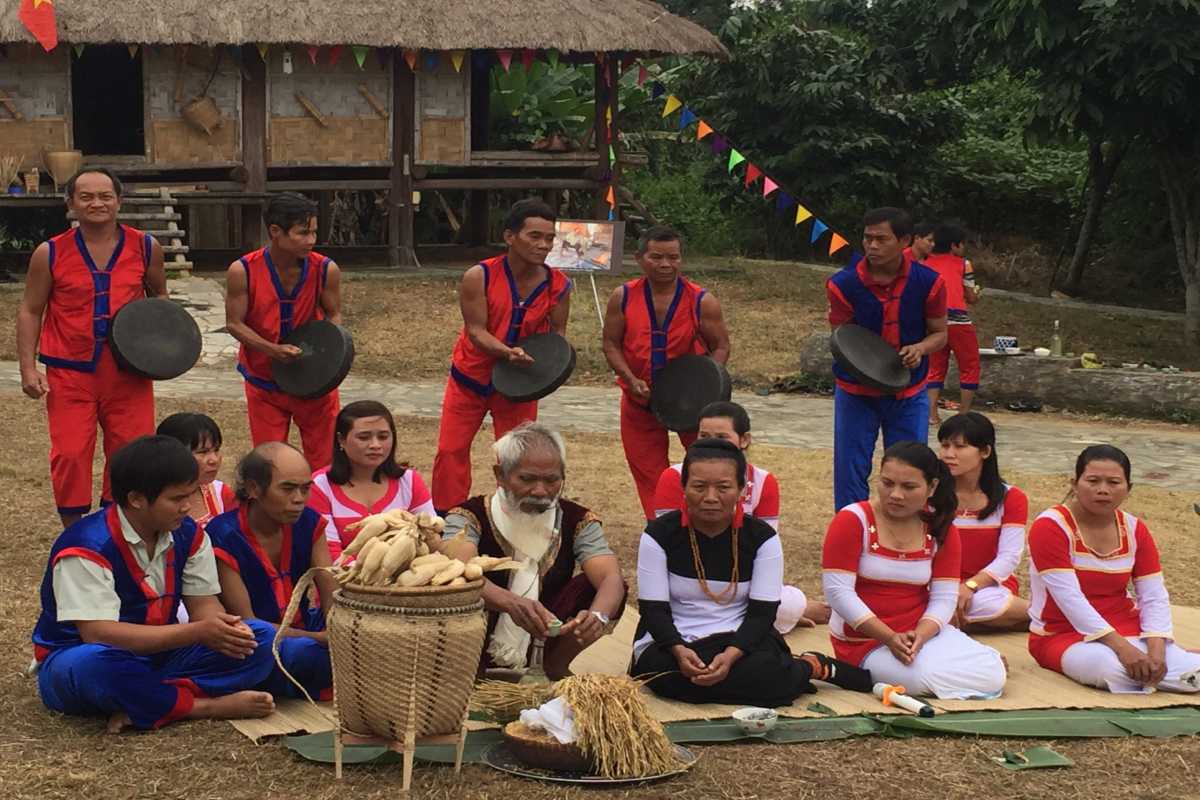 Traditional Raglai Community Gathering with Musicians Men and women in red and blue outfits gather to perform gongs during a spiritual ceremony in front of bamboo houses.