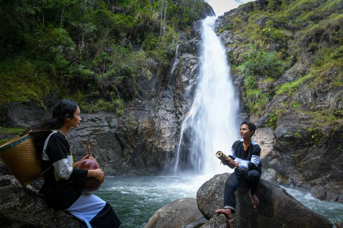 Couple in Traditional Raglai Dress at Waterfall A Raglai man and woman sit on rocks near a waterfall, holding traditional bamboo instruments during a cultural performance.