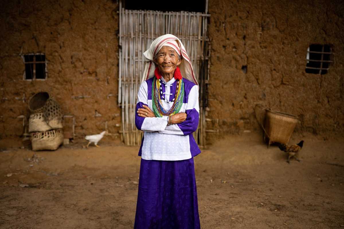 Portrait of Elderly Raglai Woman in Traditional Attire An elderly Raglai woman in purple clothing and tribal jewelry poses in front of her earthen house with a woven basket nearby.