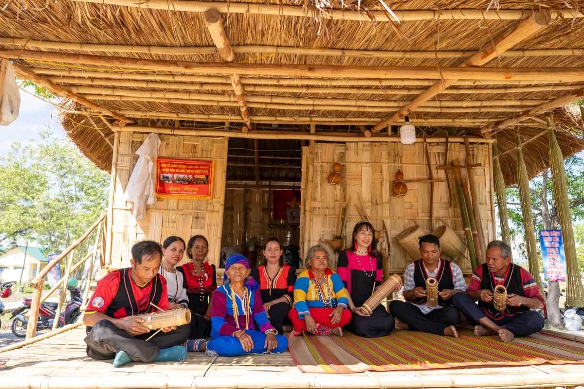 Raglai Elders and Women at Cultural House Raglai elders and women sit on a bamboo platform in front of a traditional stilt house, showcasing musical instruments and handmade goods.