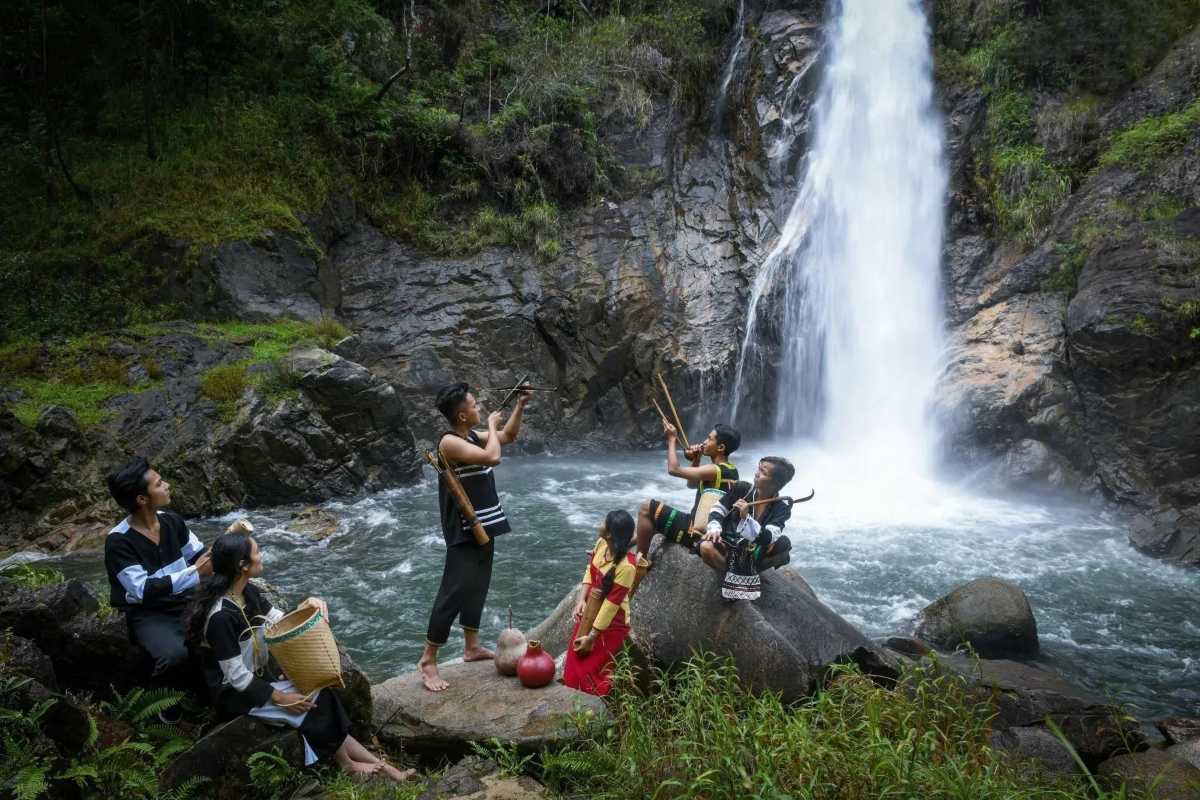 Raglai Youth Playing Music at Waterfall Raglai boys and girls dressed in tribal black outfits perform traditional flutes near a waterfall in a forested area.