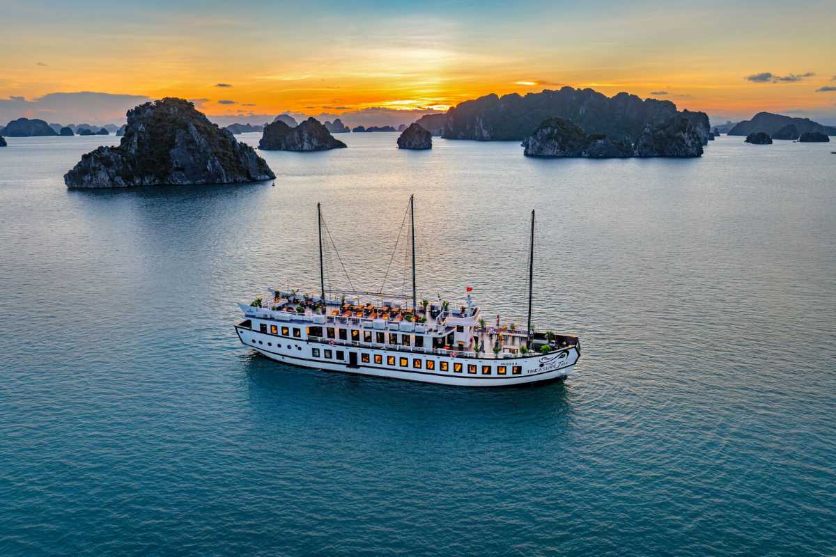 Silhouetted cruise ships sail under a dramatic sunset sky in Halong Bay, framed by lush limestone cliffs.