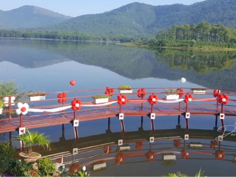 Serene Morning at Yen Trung Lake with Decorated Bridge A peaceful scene of Yen Trung Lake, featuring a red and white decorated wooden bridge over calm water, with lush green hills and forests in the background.