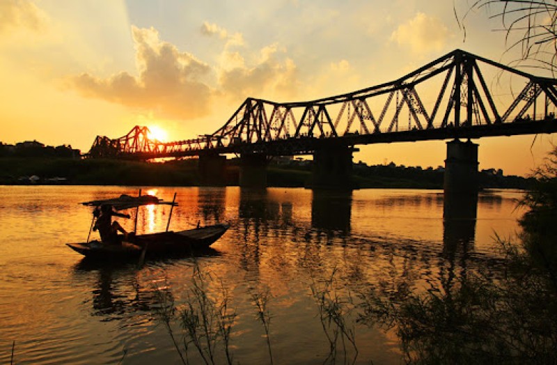 Silhouette of Long Bien Bridge at Sunset A silhouette of Long Bien Bridge at sunset with a glowing orange sky reflected on the calm waters of the Red River.