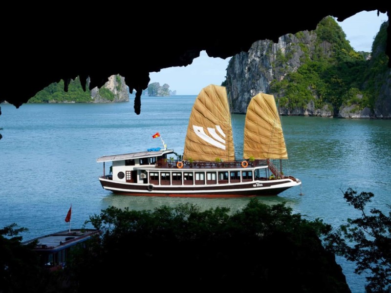 Traditional boat sailing past Bo Nau Cave in Halong Bay A traditional boat with brown sails sailing past Bo Nau Cave in Halong Bay, framed by the cave's dark rock formations.