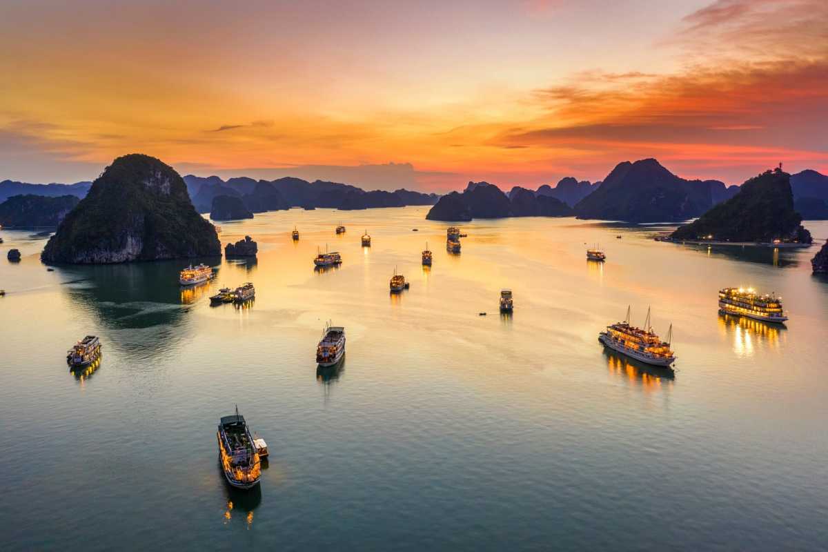 A traditional cruise boat with orange sails glides through green waters of Halong Bay during sunset.