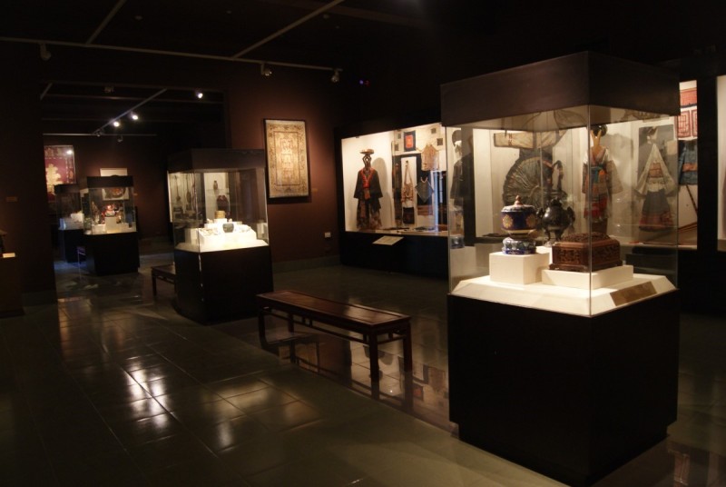 Vietnam National Fine Arts Museum Traditional Artifacts Display Dimly lit exhibit room inside Vietnam National Fine Arts Museum featuring traditional Vietnamese artifacts, ceramics, textiles, and costumes in glass cases.