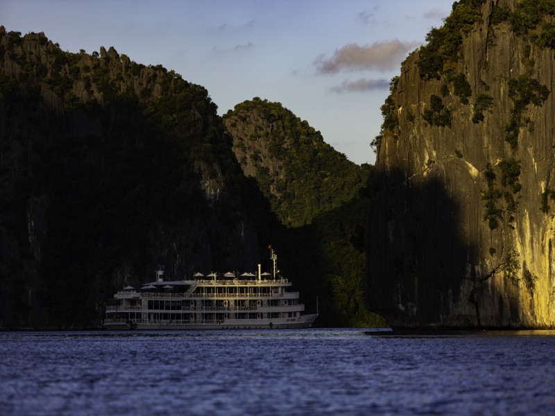 White Au Co Cruise gliding between tall limestone cliffs at golden hour in Ha Long Bay.