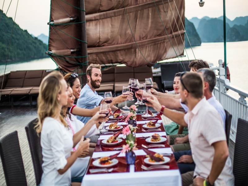 Friends toasting wine during an outdoor BBQ on Bhaya Legend Premium Cruise with Halong Bay backdrop