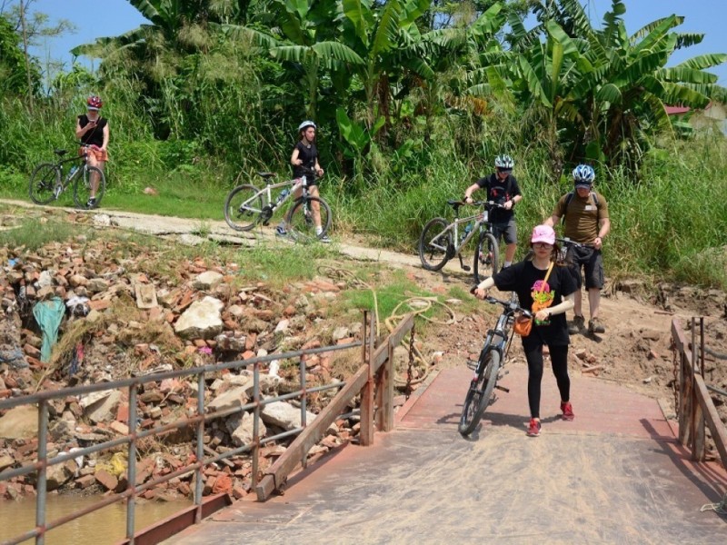 A group of cyclists crossing a small rustic bridge in Vietnam amidst lush greenery.